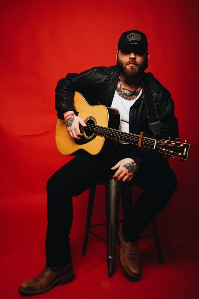 East Rich sitting on a stool, holding an acoustic guitar, in front of a deep red background.